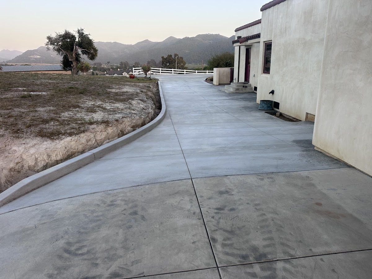 A wide concrete driveway curving past a stucco house, with a poured curb following the cut bank and the Arroyo Grande hills behind at dusk.